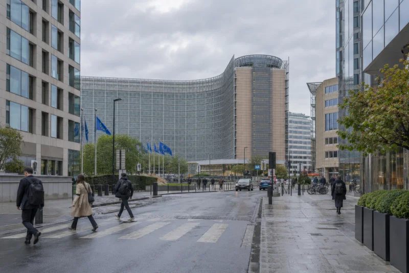 Transeúntes caminan por una calle del barrio europeo de Bruselas, entre edificios de oficinas bajo un cielo nublado.