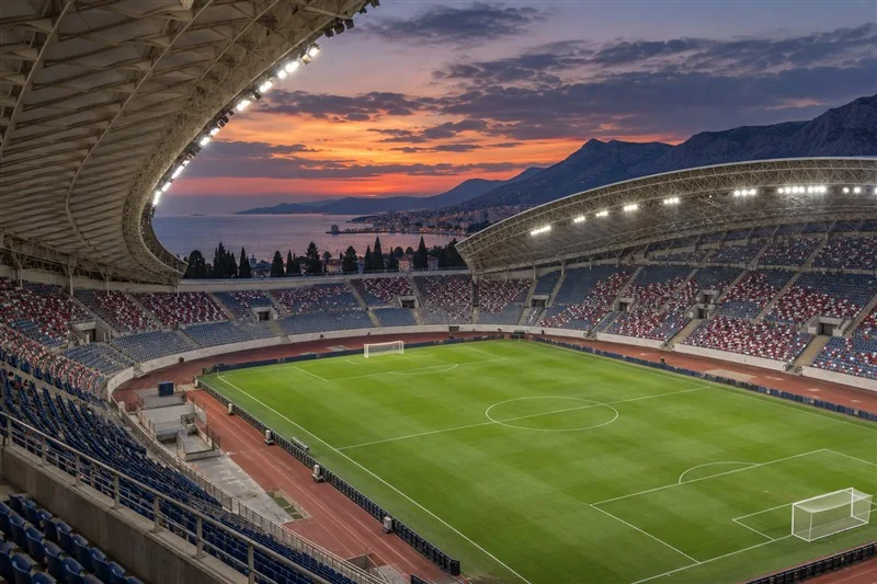 Vista de un campo de fútbol vacío al atardecer, con cubierta blanca y gradas azules.